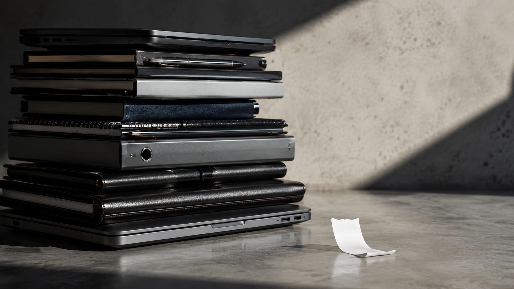 Must visibly show: A large stack of office supplies next to a tiny receipt. Local anchors: compra de material, recibo simple, carece de validez. Style family: conceptual-still-life. Visual brief: A towering stack of premium office equipment, including a closed laptop, leather portfolios, and sleek metallic pens, resting on a polished concrete floor. Beside this expensive pile sits a single, tiny, crumpled strip of blank thermal paper, visually emphasizing the inadequacy of a simple receipt for large purchases. Dramatic studio lighting with a strong directional key light casts deep shadows, highlighting the disparity in scale. Shot on a Fujifilm GFX 100S with a 63mm lens. Editorial still life photography. Inline body image text firewall: visual content only. Do not render readable words anywhere. No article title, headline, subtitle, cover line, typography lockup, title card, poster text, labels, badges, UI copy, logo, brand name, or wordmark. If an interface, document, workflow, chart, or screen is needed, show abstract unreadable marks, shapes, icons, lines, and color blocks instead of words. Avoid repeating: police or inspectors, gavel, handcuffs.